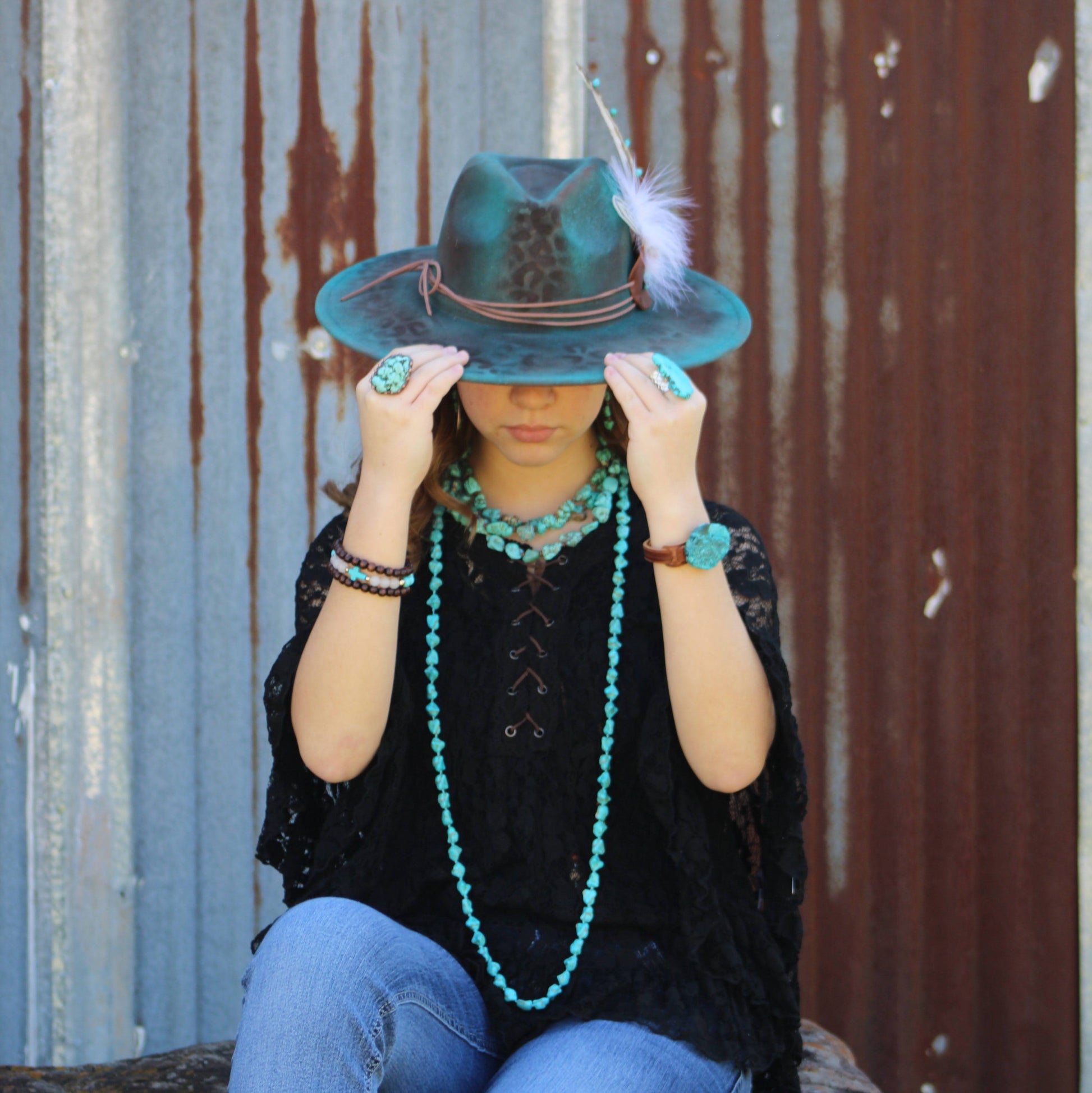 Person wearing a turquoise hat with a feather, black top, and blue jeans, sitting against a rusty metal wall.