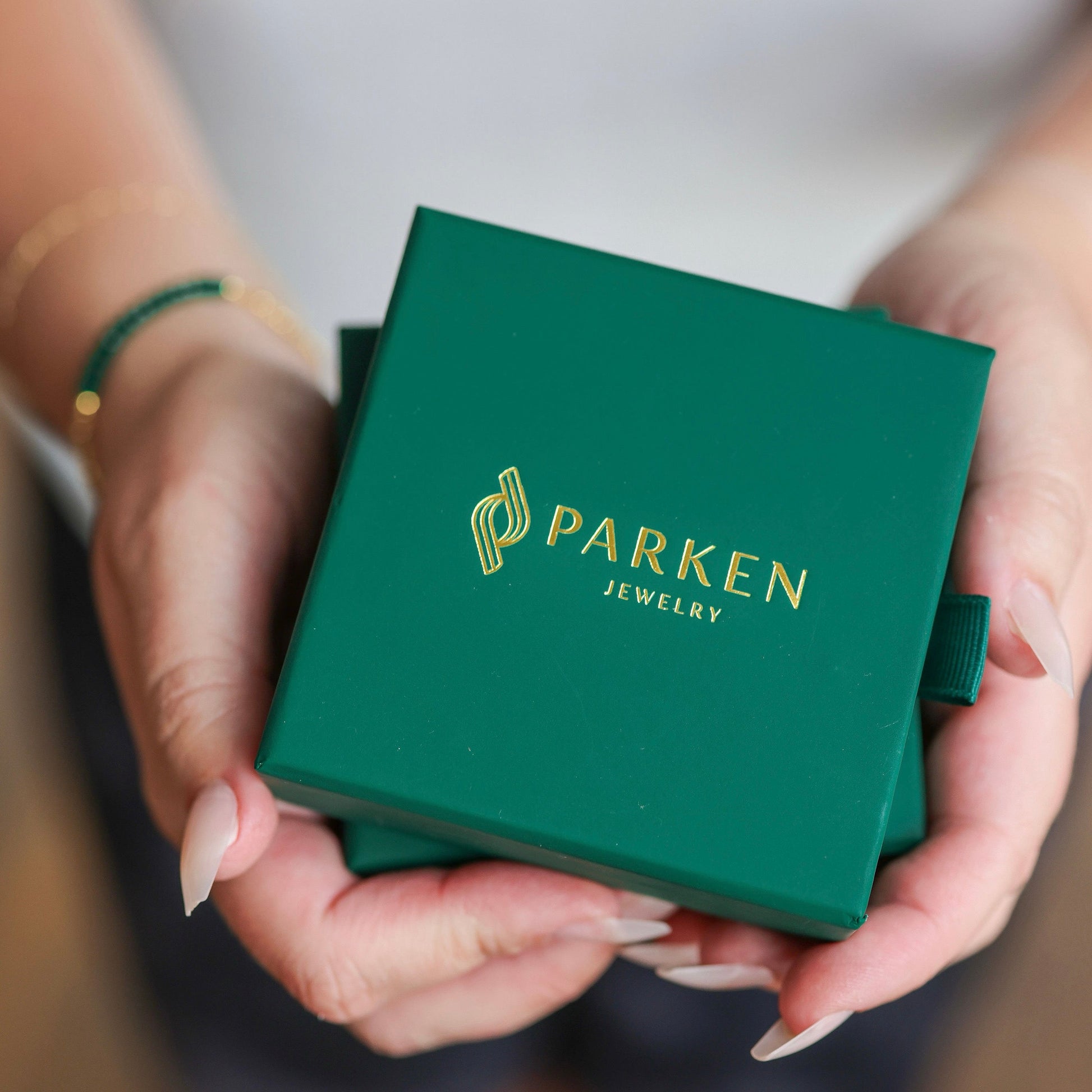 Hands holding a square green jewelry box with gold PARKEN JEWELRY text printed on the lid.