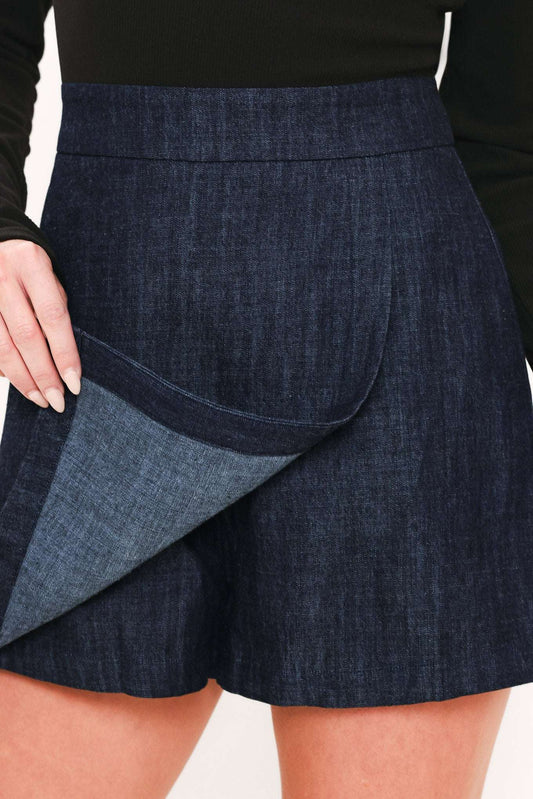 Close-up of a model wearing dark blue denim shorts with a matching denim lining shown being pulled up, paired with a black long-sleeve top.