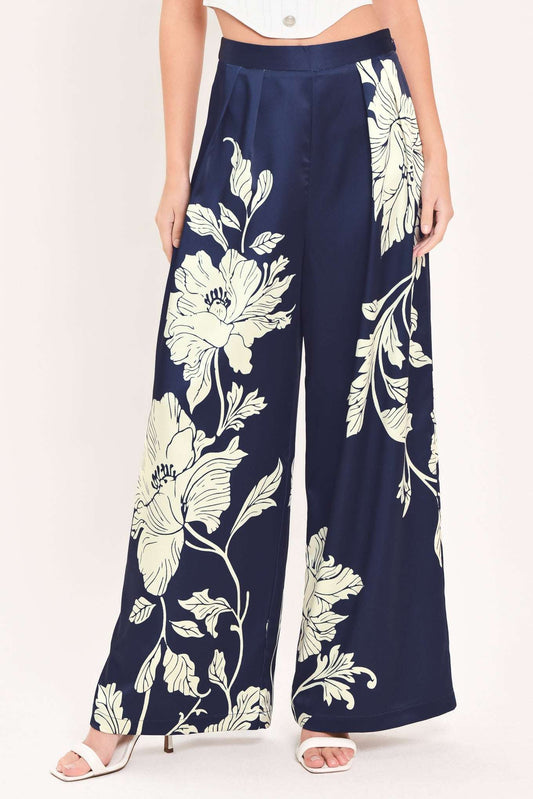 A model wearing navy blue wide-leg pants with a large cream floral print, paired with a white crop top and white heeled sandals, against a plain white background.