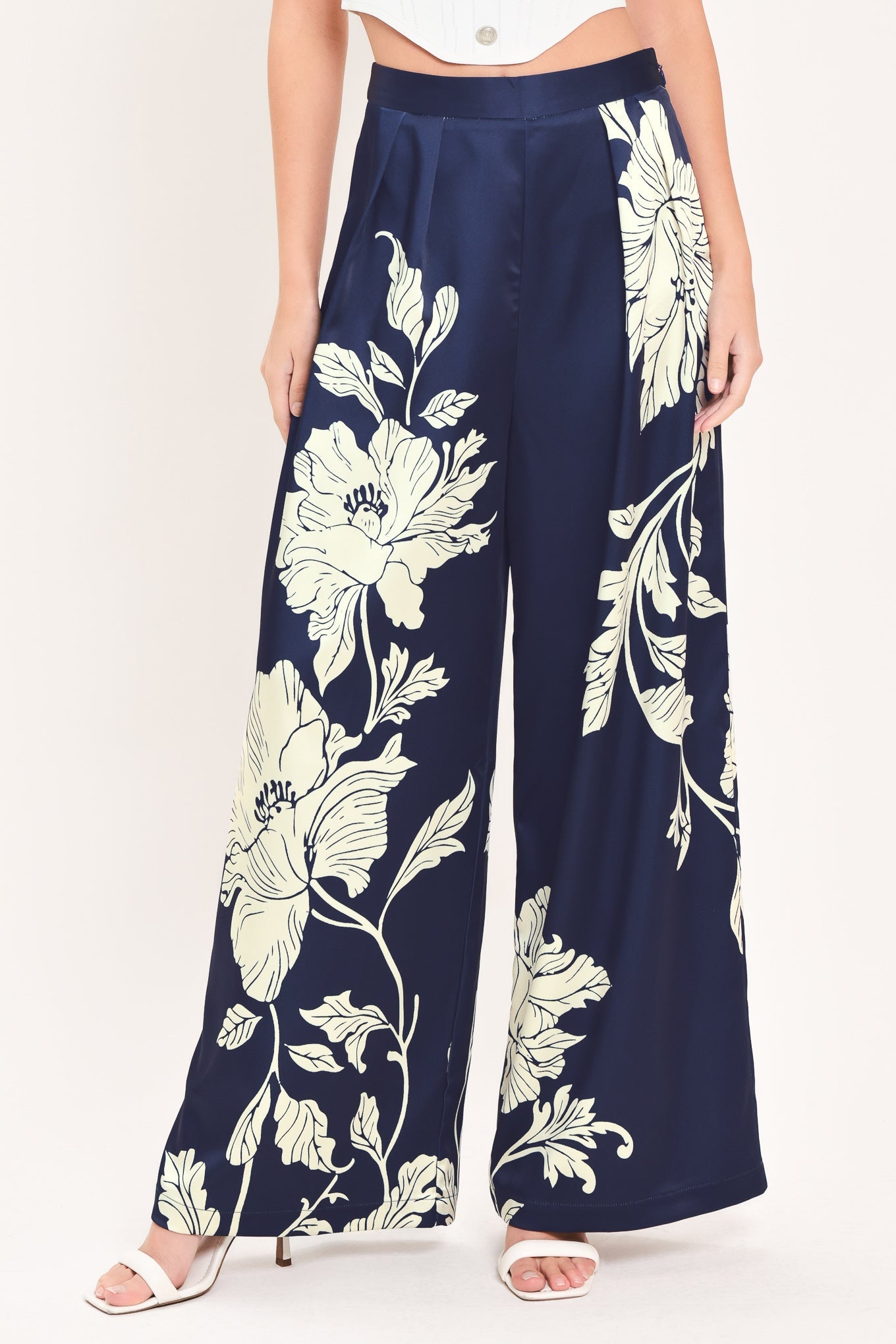 A model wearing navy blue wide-leg pants with a large cream floral print, paired with a white crop top and white heeled sandals, against a plain white background.