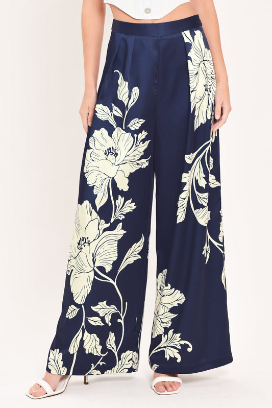 A model wearing navy blue wide-leg pants with a large cream floral print, paired with a white crop top and white heeled sandals, against a plain white background.