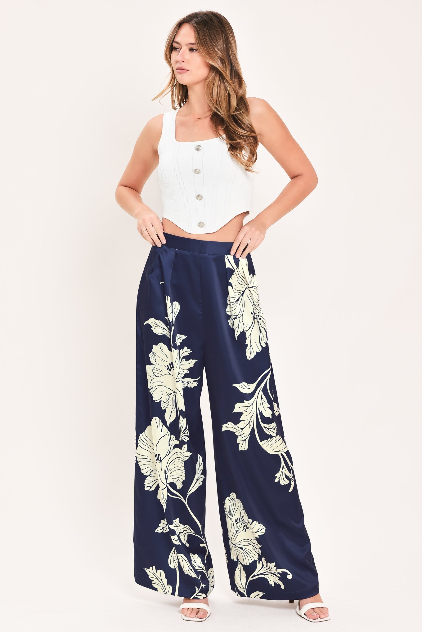 A woman modeling a pair of navy blue wide-leg pants with a large white floral print, paired with a white sleeveless crop top, against a plain white studio background.