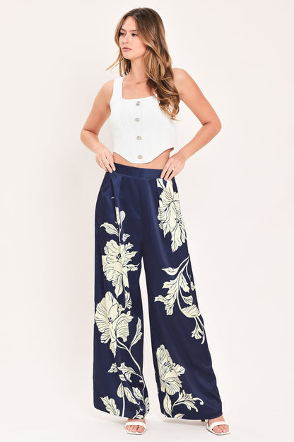 A woman modeling a pair of navy blue wide-leg pants with a large white floral print, paired with a white sleeveless crop top, against a plain white studio background.