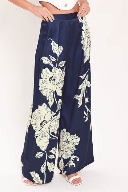 A person wearing wide-leg navy pants with a large cream floral print, paired with a white cropped top and white heeled sandals, against a plain light background.