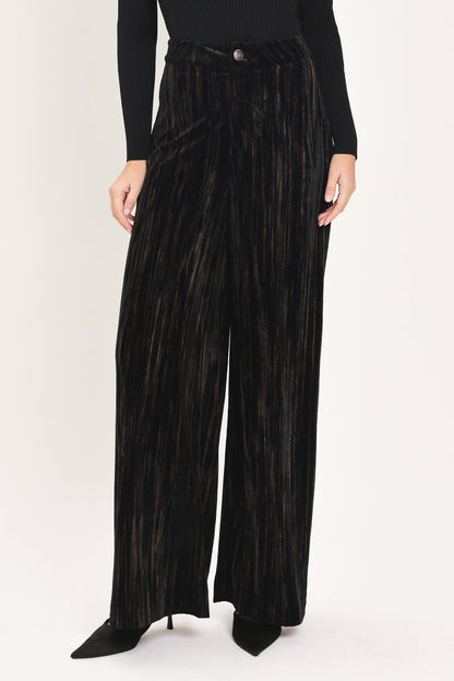 A model wearing high-waisted, wide-leg black pleated velvet trousers with a button waistband, paired with a black long-sleeve top and black pointed-toe heels, against a plain white studio background.