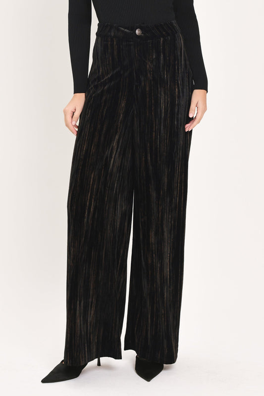 A model wearing high-waisted, wide-leg black pleated velvet trousers with a button waistband, paired with a black long-sleeve top and black pointed-toe heels, against a plain white studio background.