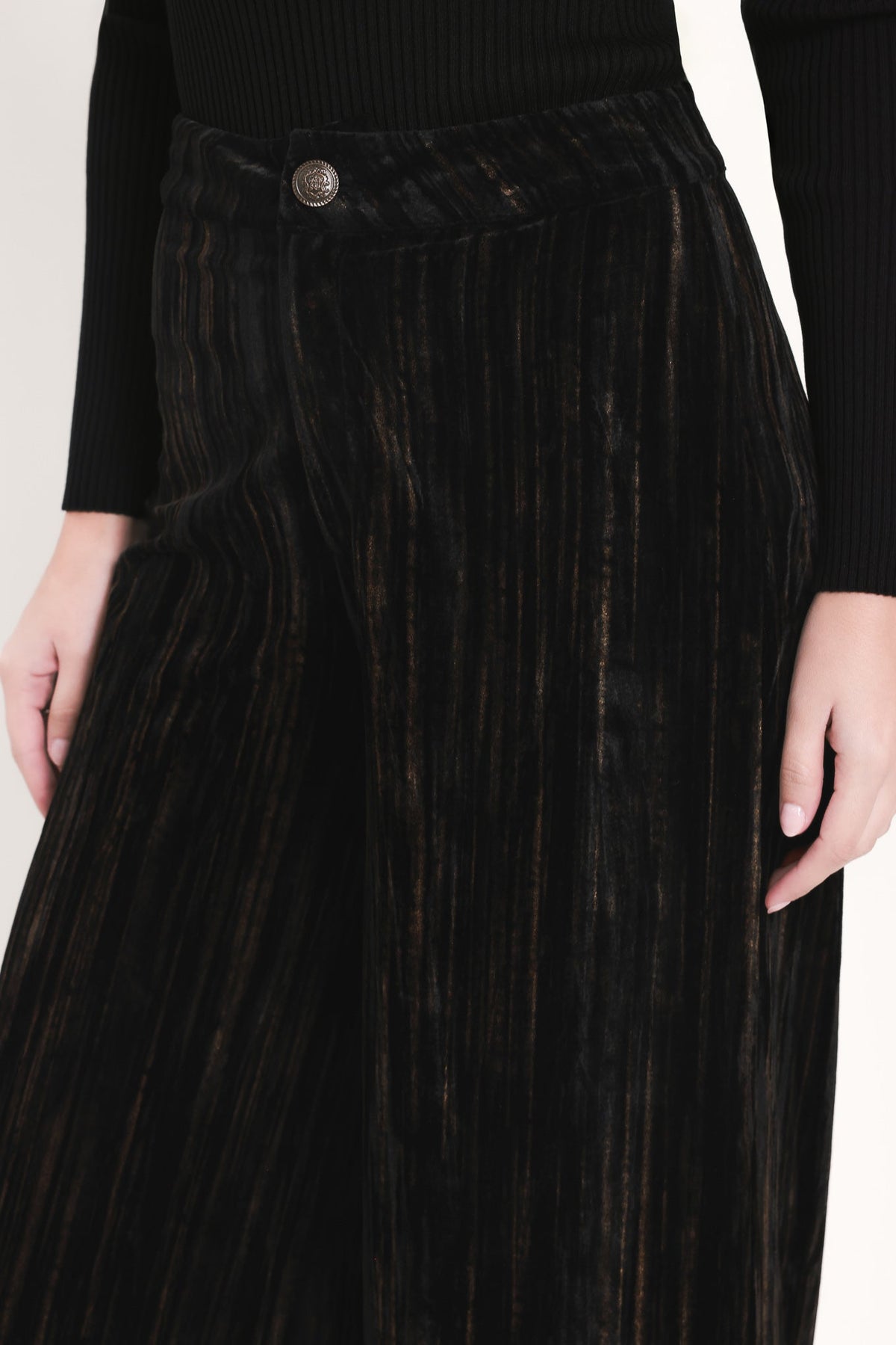 Close-up of a person wearing a black ribbed long-sleeve top and a black pleated velvet skirt with subtle warm brown undertones and a single button closure.