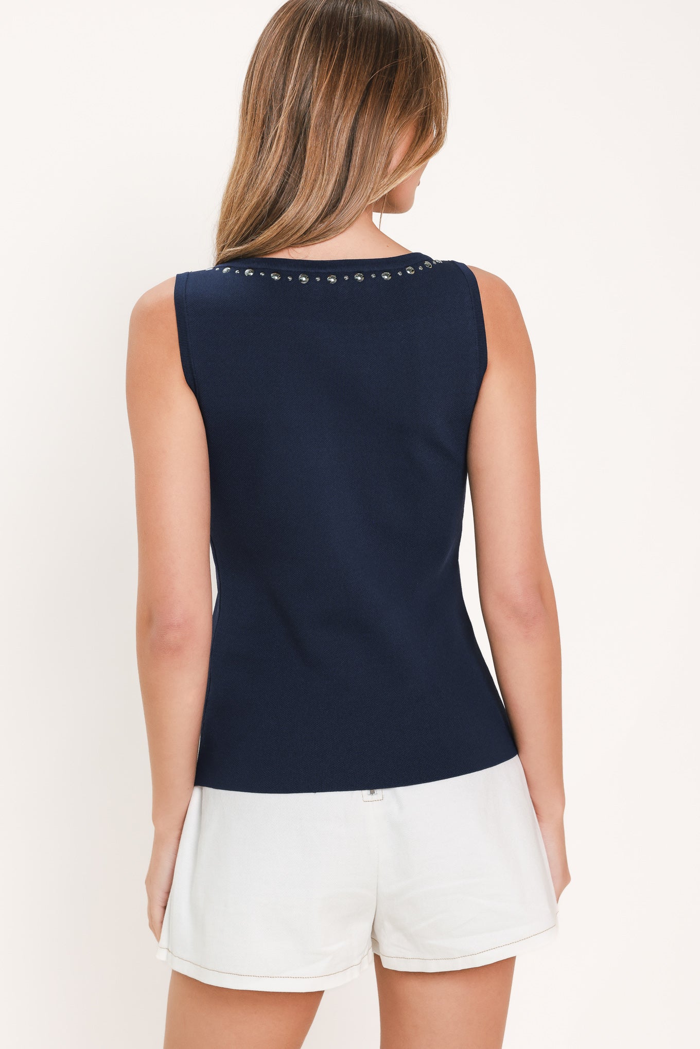 A woman modeling a sleeveless navy blue top and a white pleated skirt,  against a solid light beige background. back view.