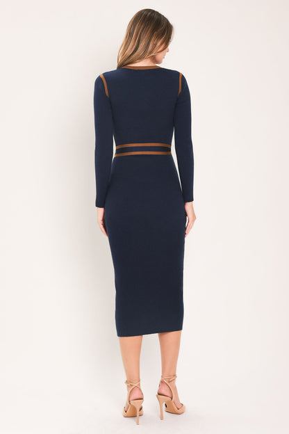 A woman viewed from the back wearing a navy midi dress with long sleeves and thin brown trim details at the shoulders and waist, paired with beige high-heeled sandals against a plain light background.