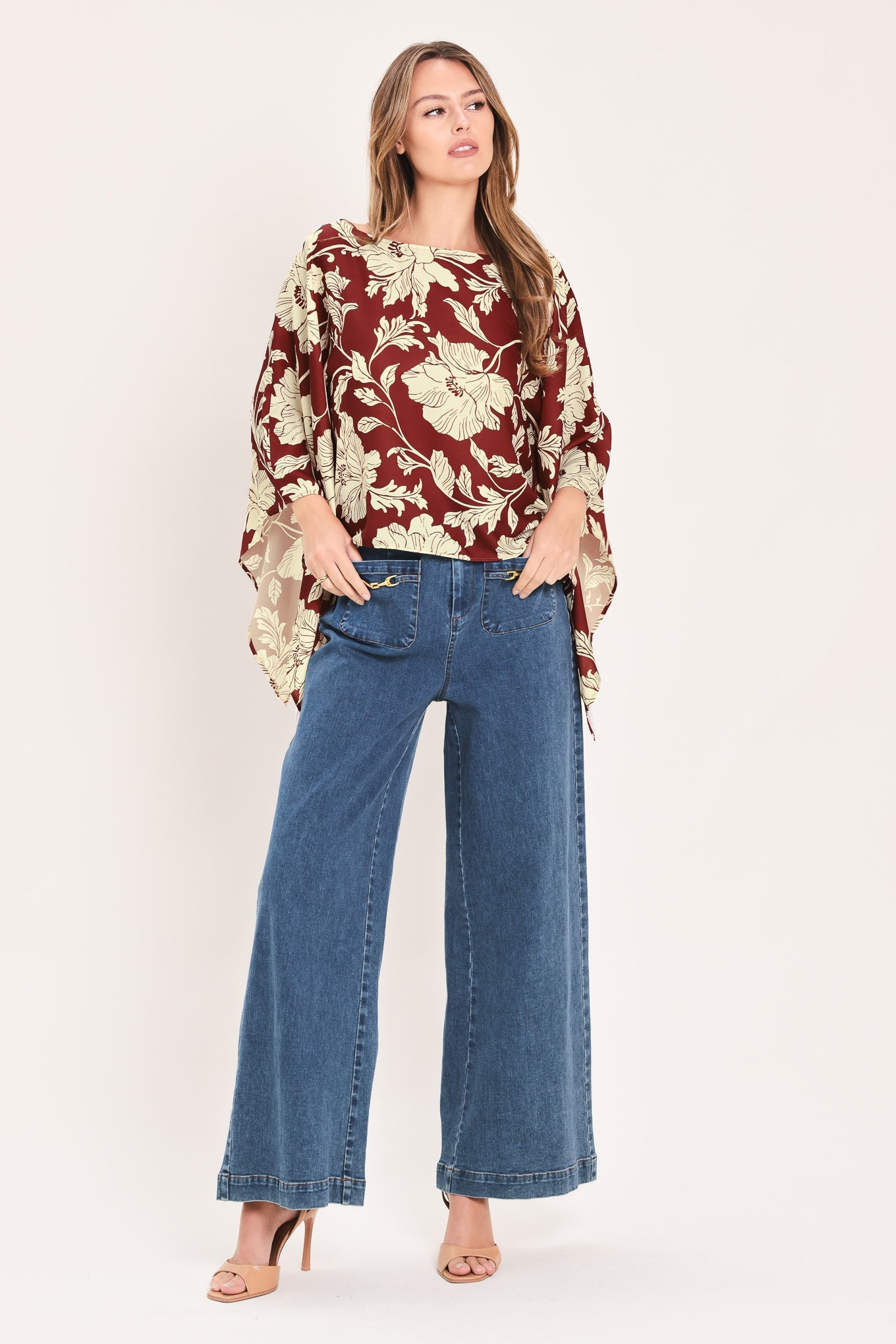 A woman modeling a rust red and beige floral print blouse with wide bell sleeves, paired with blue wide-leg jeans and nude high-heeled sandals, against a solid off-white studio background.