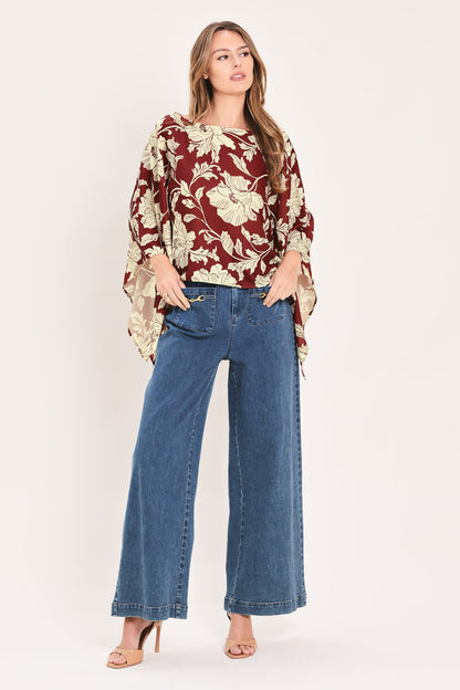 A woman modeling a rust red and beige floral print blouse with wide bell sleeves, paired with blue wide-leg jeans and nude high-heeled sandals, against a solid off-white studio background.