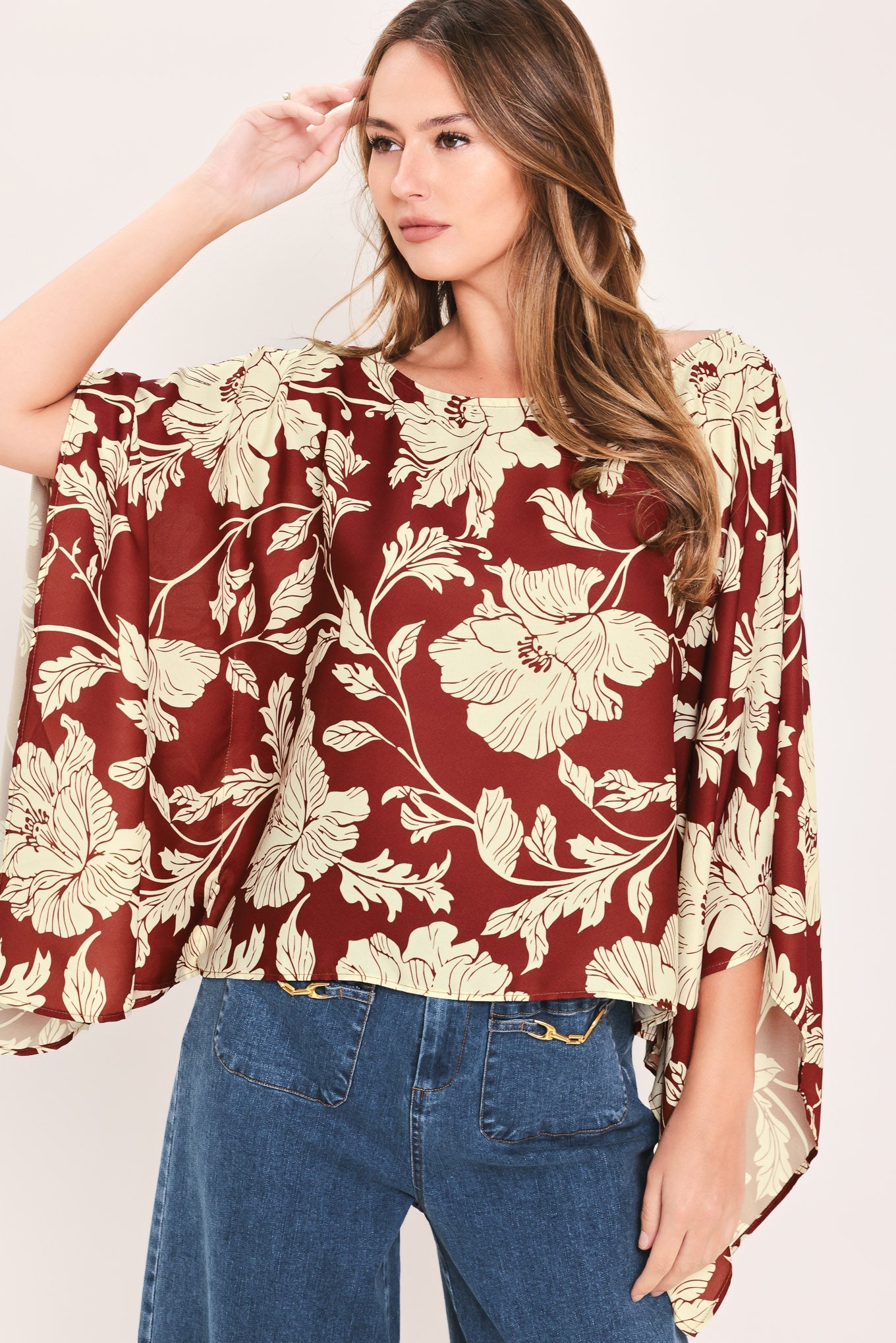A woman wearing a rust red and cream floral print top with wide bell sleeves, paired with blue jeans, against a plain light background.