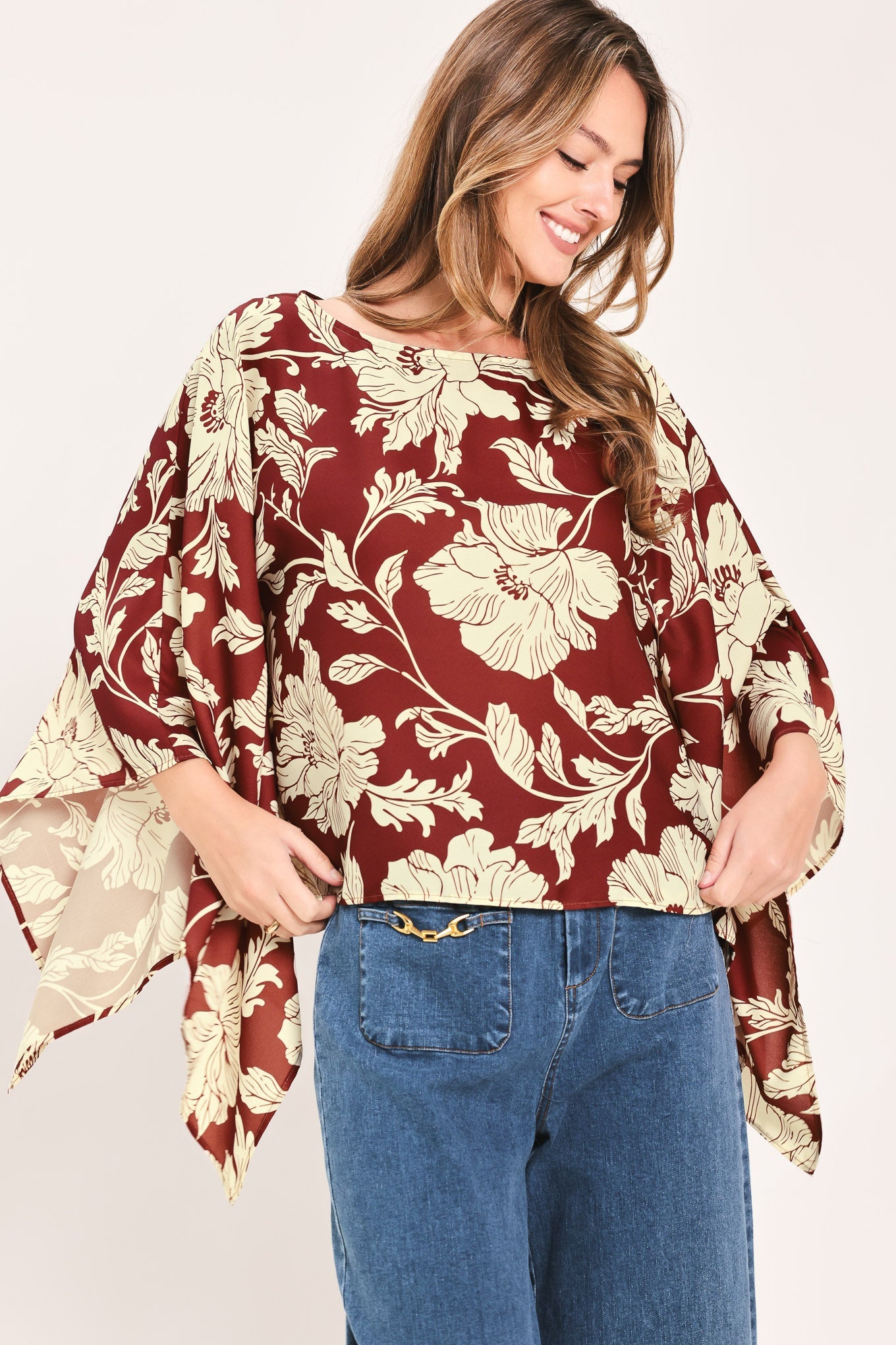 A woman wearing a loose, short-sleeve blouse with a burgundy and beige floral print, paired with blue jeans, against a plain light background.