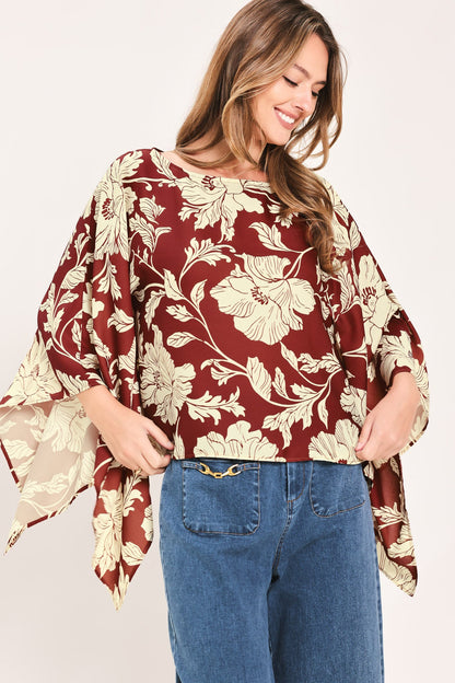A woman wearing a loose, short-sleeve blouse with a burgundy and beige floral print, paired with blue jeans, against a plain light background.