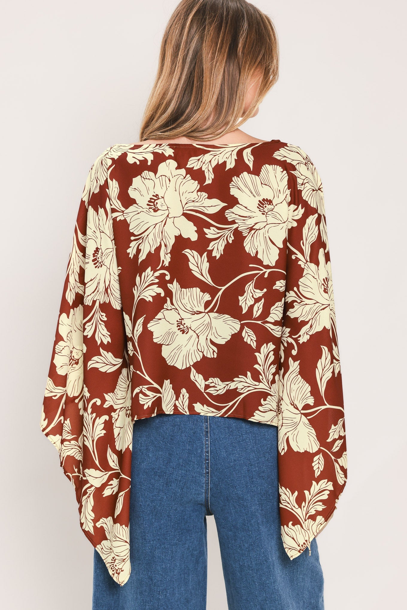 A woman viewed from the back wearing a rust-brown long-sleeve blouse with a cream floral print and blue jeans, set against a plain light gray studio background.