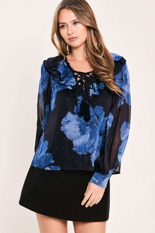 A woman modeling a sheer black blouse with a blue floral print, ruffled collar, and lace-up neckline, paired with a black skirt, against a plain light background.