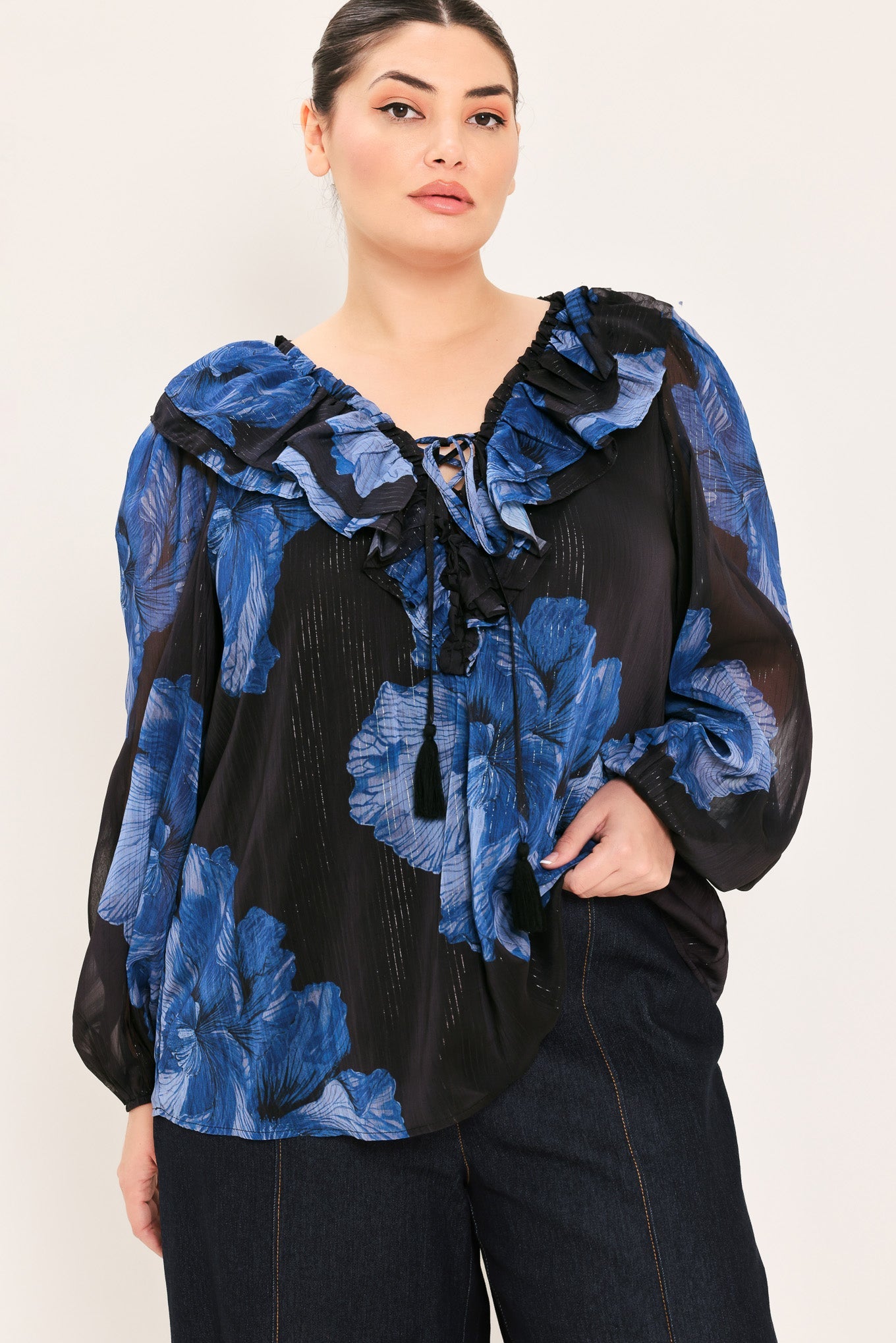 A woman wearing a black and blue floral long-sleeve blouse with ruffled details, paired with dark jeans, against a plain light background.