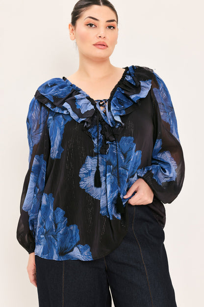A woman wearing a black and blue floral long-sleeve blouse with ruffled details, paired with dark jeans, against a plain light background.