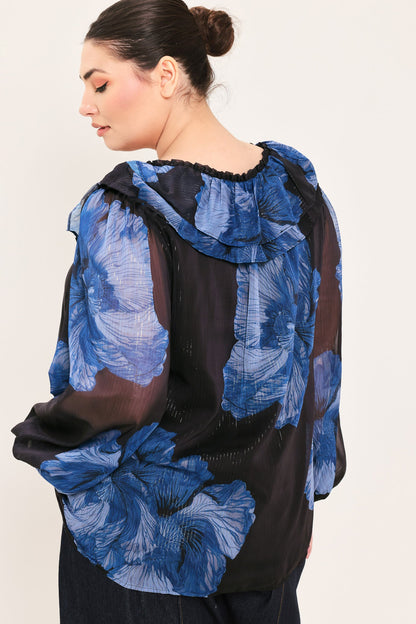 A woman shown from the back wearing a sheer black blouse with large blue floral print and ruffled detailing, against a plain light background.