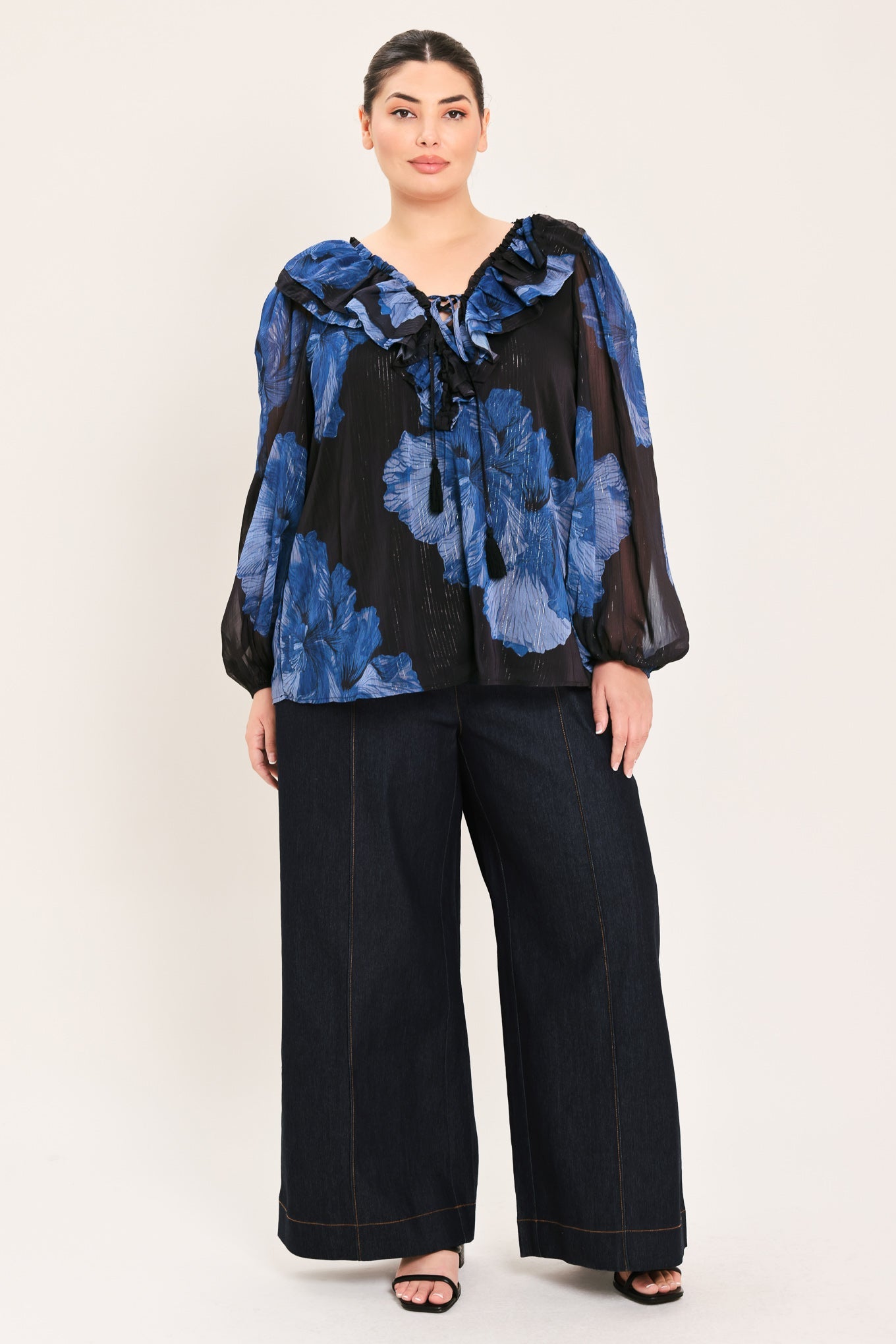 A woman modeling a sheer black long-sleeve blouse with a blue floral print and wide-leg dark pants against a plain light beige studio background.