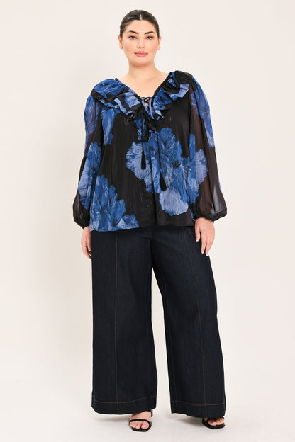A woman modeling a sheer black long-sleeve blouse with a blue floral print and wide-leg dark pants against a plain light beige studio background.