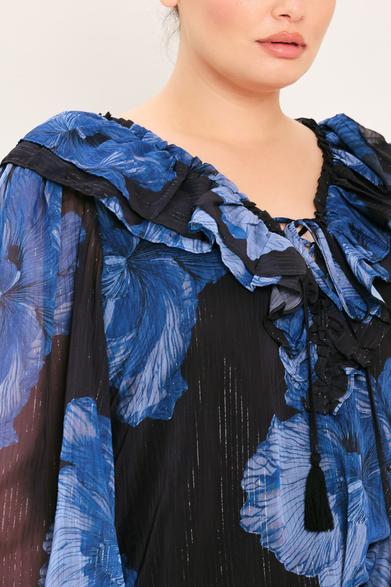 Close-up of a model wearing a sheer black and blue floral print top with dramatic ruffled shoulders and a tassel tie at the neckline.