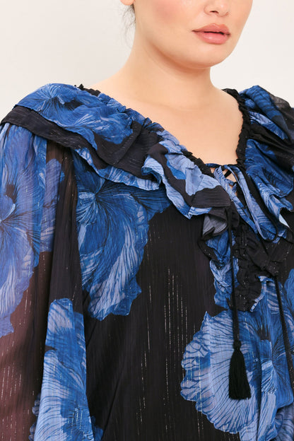 Close-up of a model wearing a sheer black and blue floral print top with dramatic ruffled shoulders and a tassel tie at the neckline.