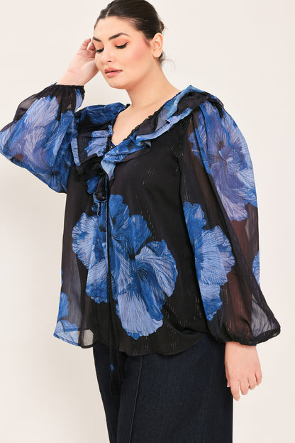 A woman wearing a sheer black blouse with large blue floral print and ruffled long sleeves, paired with dark pants, against a plain light background.