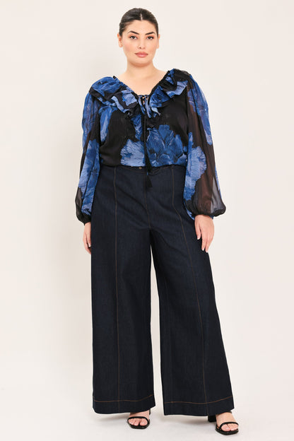 A woman modeling a sheer black and blue floral long-sleeve blouse with a ruffled collar and front tie, paired with dark wide-leg pants, against a plain light beige studio background.