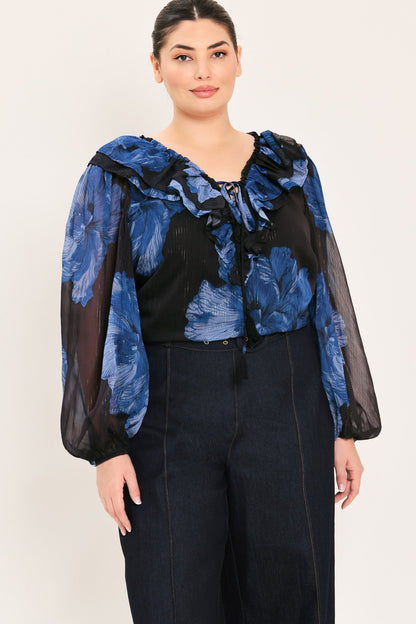 A woman modeling a sheer black blouse with a blue floral print and ruffled collar, paired with dark wide-leg pants, against a plain light background.