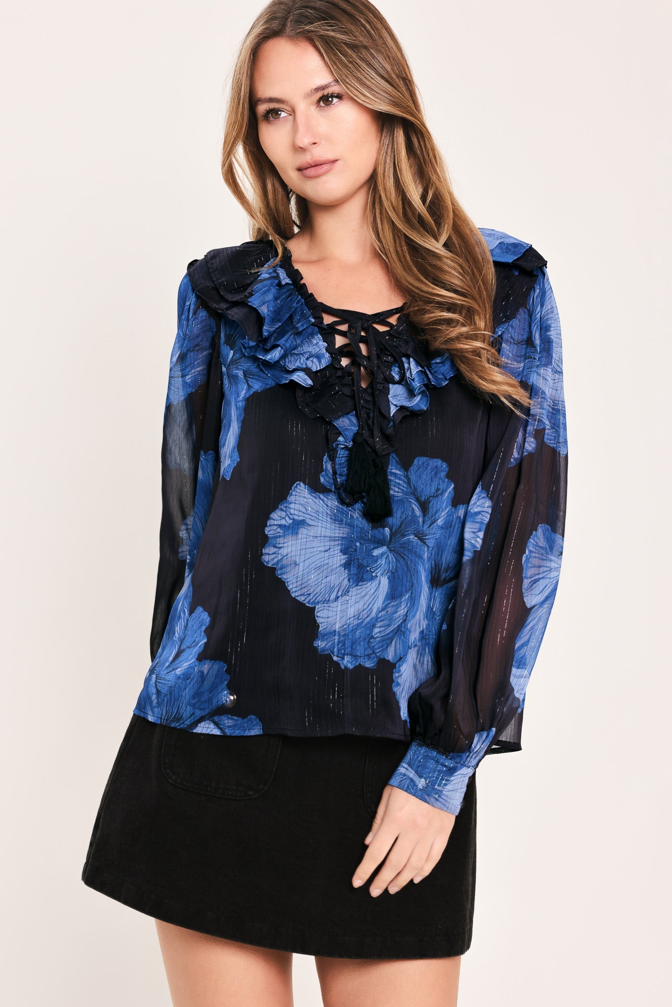 A woman modeling a sheer black blouse with a blue floral print, ruffled collar, and lace-up neckline, paired with a black skirt, against a plain light background.