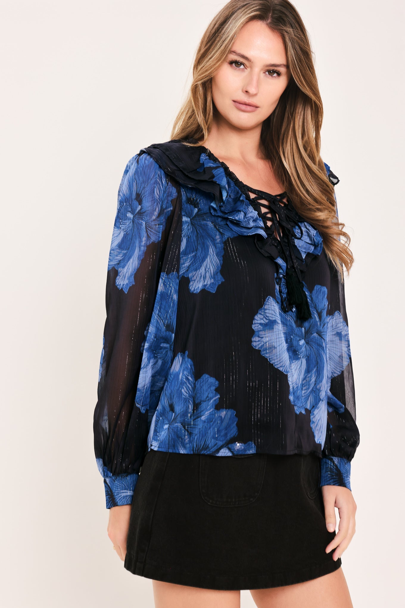 A woman wearing a black and blue floral long-sleeve blouse with a ruffled collar and lace-up neckline, paired with a black skirt, against a plain light background.