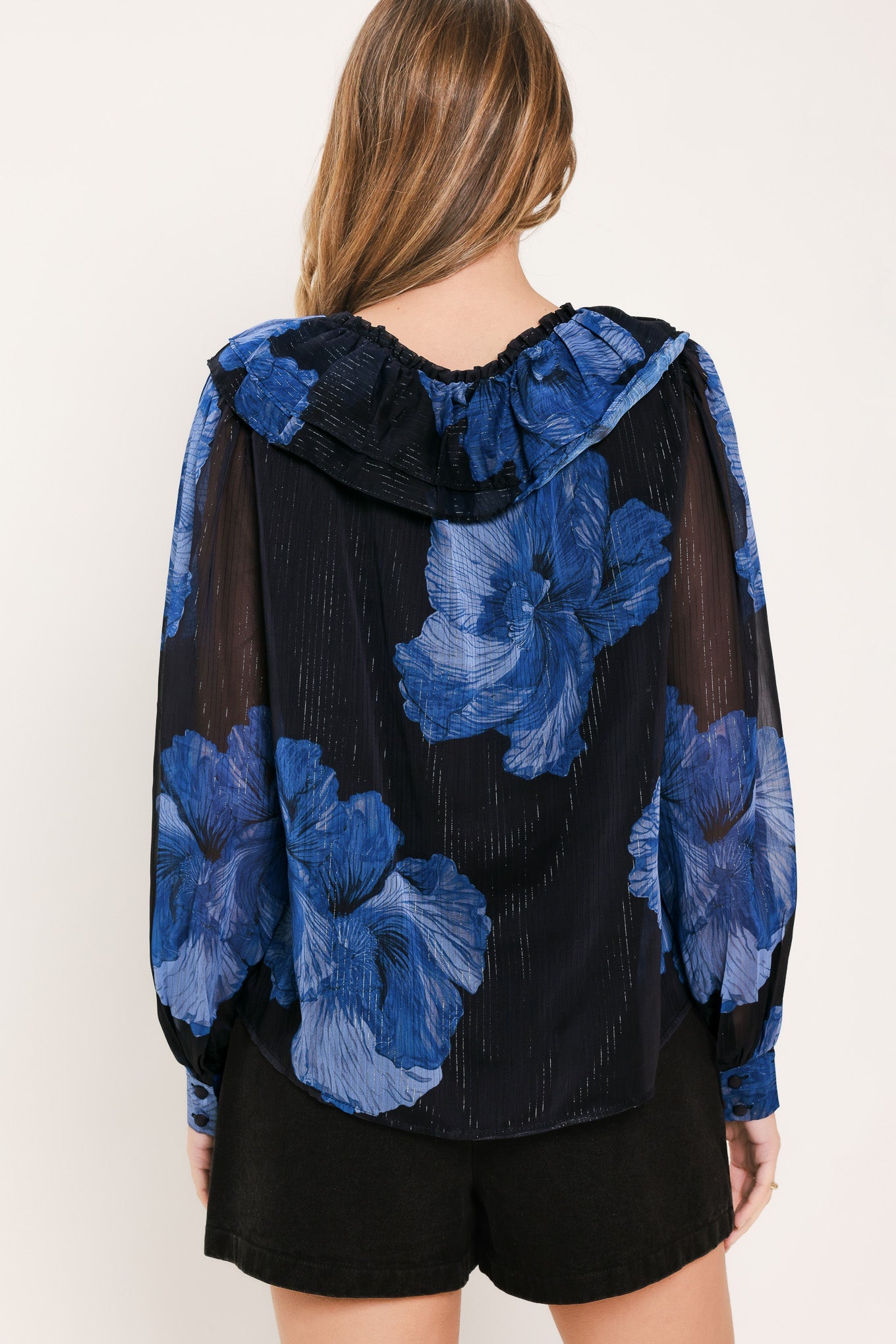 A woman shown from the back wearing a sheer black long-sleeve top with a large blue floral print and a ruffled neckline, paired with black shorts against a plain light background.
