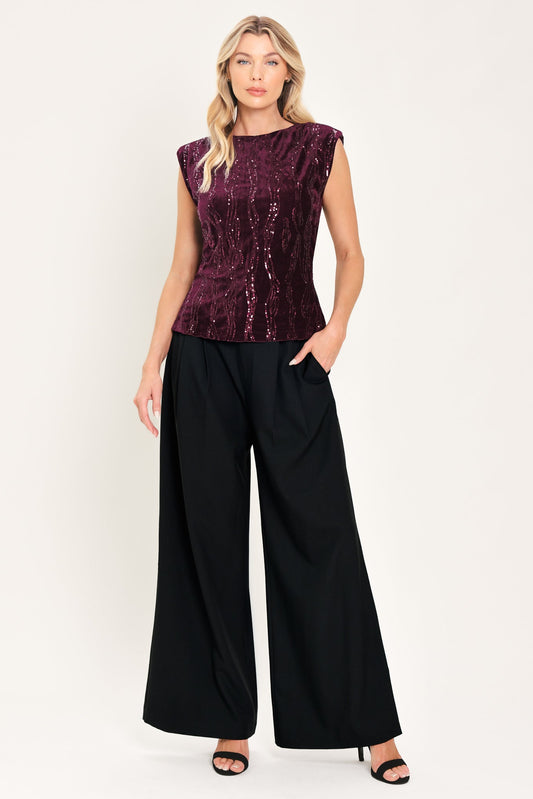 A woman modeling a sleeveless deep plum sequined top paired with wide-leg black pants, standing against a plain light background.