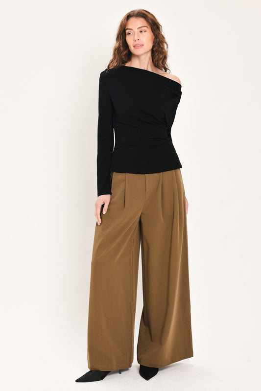 A woman modeling a black off-the-shoulder long-sleeve top paired with wide-leg brown trousers, standing against a plain light background.
