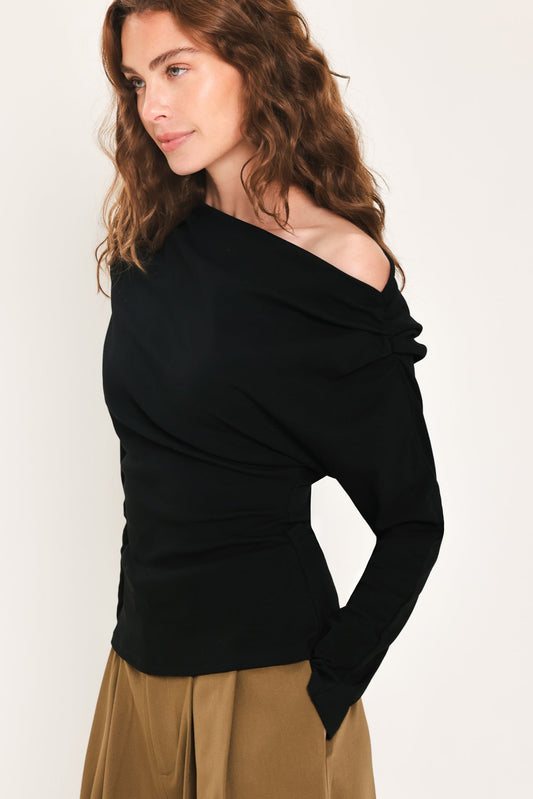 A woman wearing a black off-the-shoulder long-sleeve knit top and tan skirt, photographed against a plain light background.