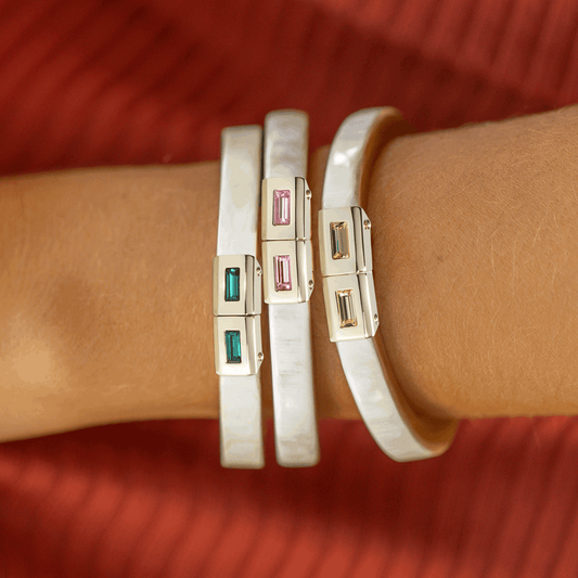 Close-up of a wrist wearing two white bangles with gold and gemstone accents on a red background.