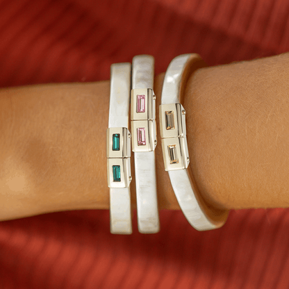 Close-up of a wrist wearing two white bangles with gold and gemstone accents on a red background.