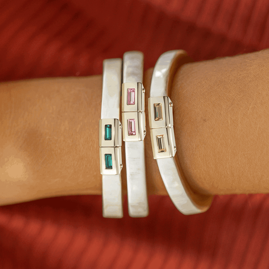 Close-up of a wrist wearing two white bangles with gold and gemstone accents on a red background.