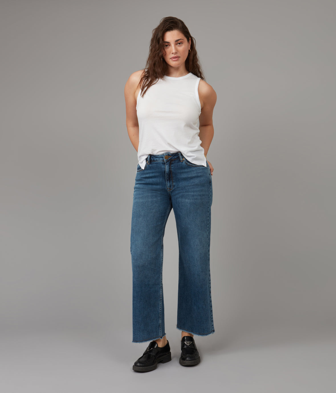 Woman wearing a white tank top and blue jeans against a gray background