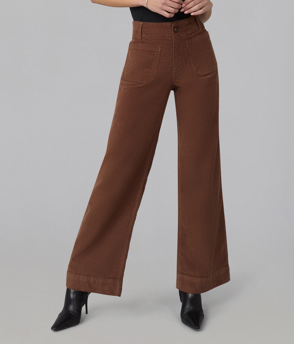 A person wearing high-waisted, wide-leg brown pants with black pointed-toe heeled boots, shown from the waist down against a plain light gray studio background.