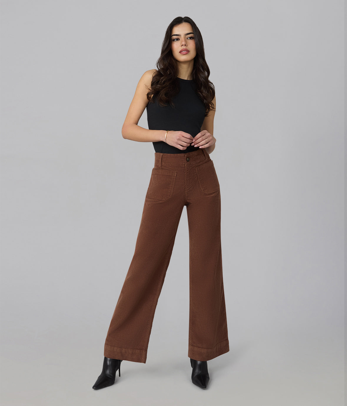 A woman modeling a pair of high-waisted, wide-leg brown pants with a black sleeveless top and black pointed-toe heels, standing against a solid light gray studio background.
