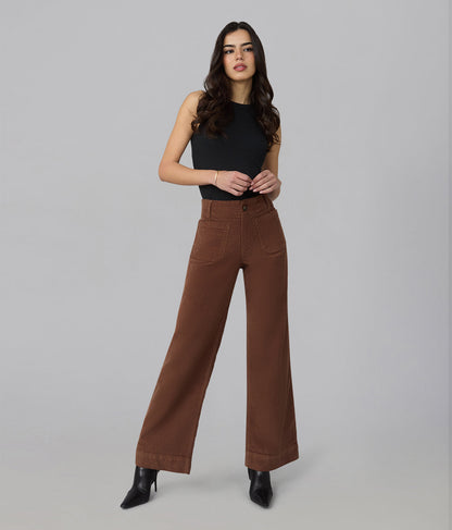 A woman modeling a pair of high-waisted, wide-leg brown pants with a black sleeveless top and black pointed-toe heels, standing against a solid light gray studio background.