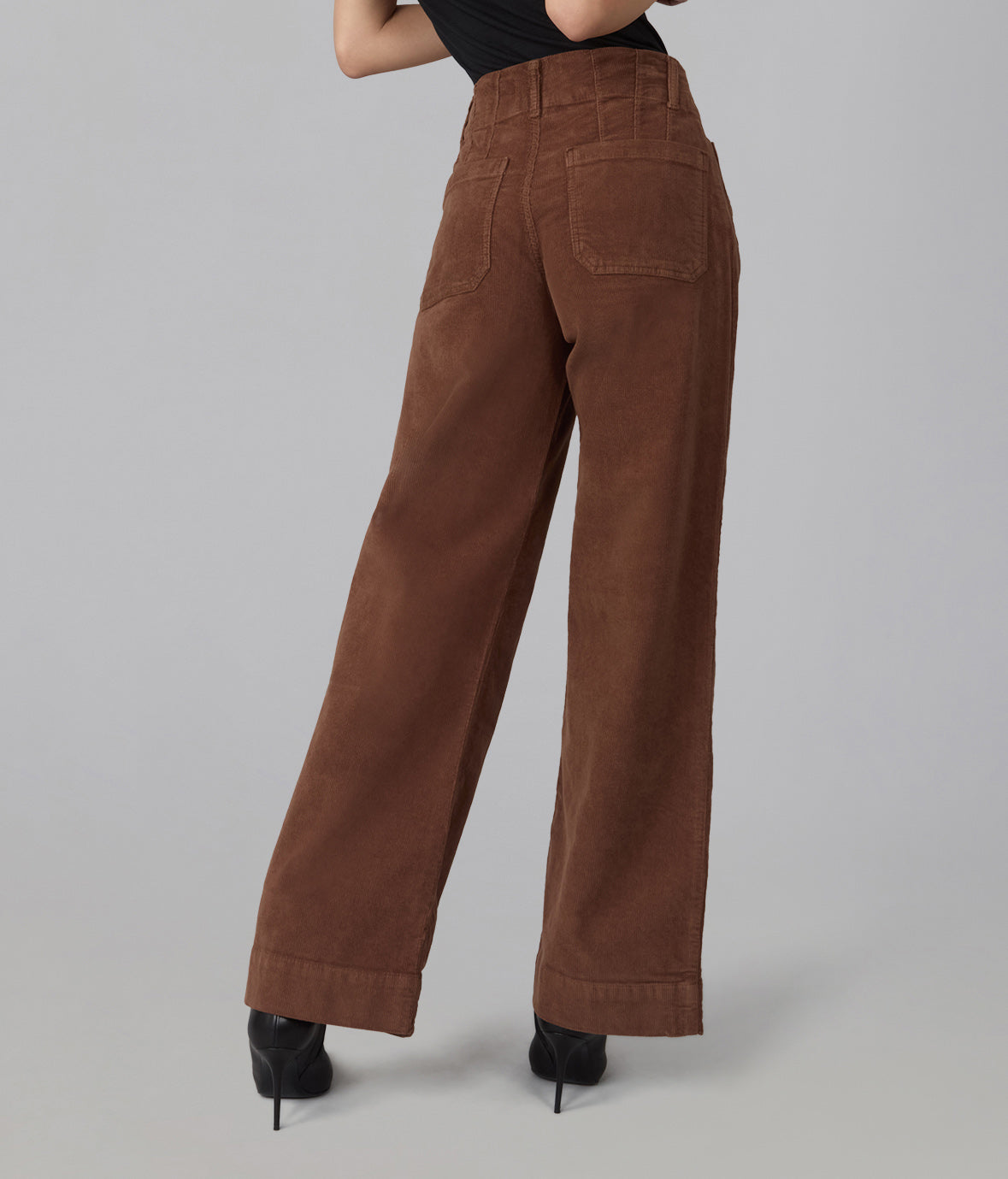 A person shown from the back wearing high-waisted, wide-leg brown corduroy pants with black high heels against a plain light gray studio background.