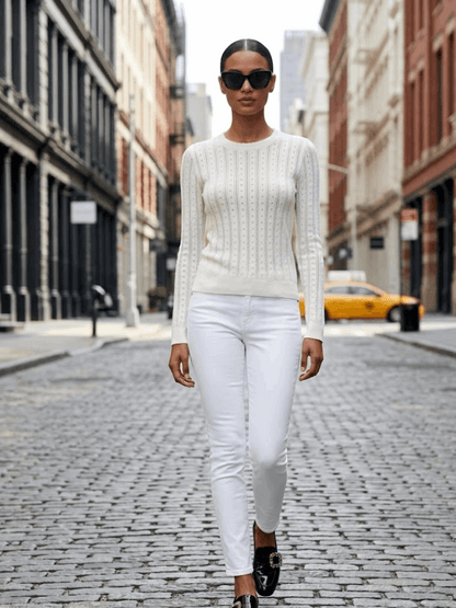Model wearing a white pointelle knit sweater in white with white jeans and a city street background.