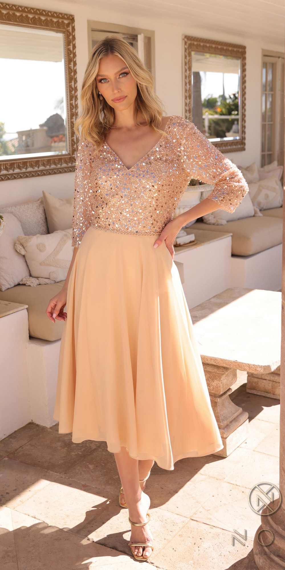 Woman in a beige dress with sequin top standing in a sunlit room.