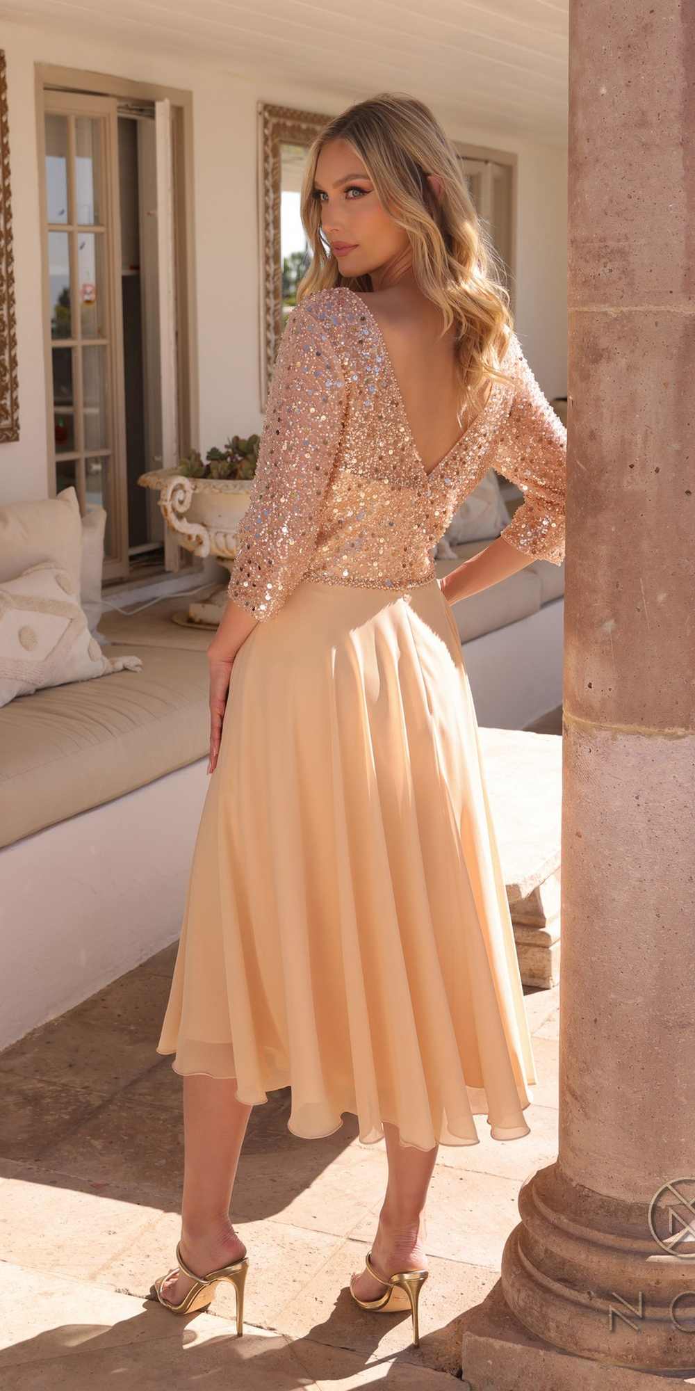 Woman in a sparkling top and flowing skirt standing in a sunlit room with columns.