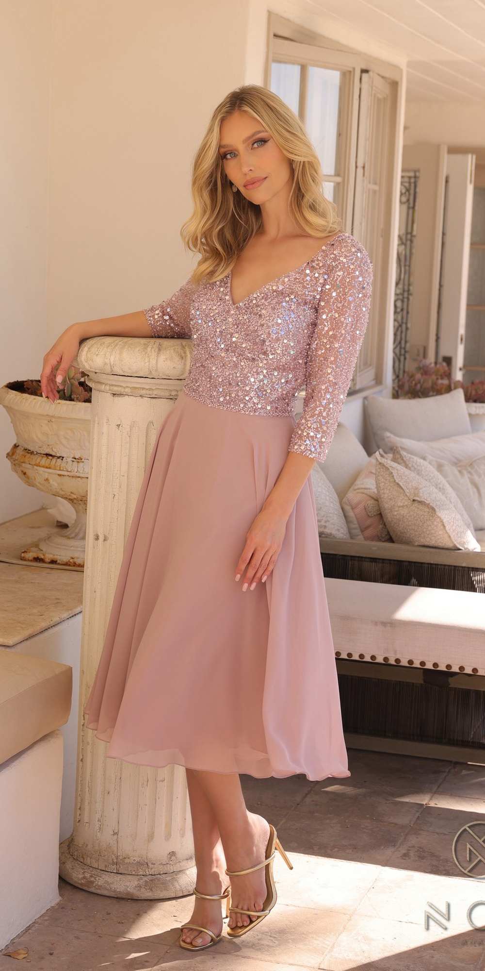 Woman in a pink dress standing in a sunlit room with large windows.