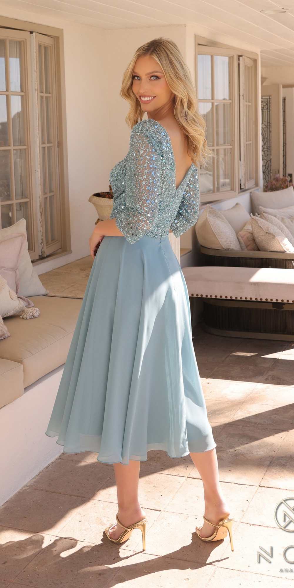 Woman in a light blue dress with a beaded top standing in a sunlit room.